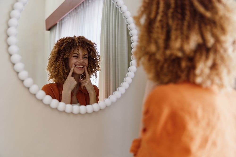 Femme souriante aux cheveux bouclés devant son miroir Femme aux cheveux bouclés souriant dans un miroir perlé, pointant ses fossettes avec ses doigts.