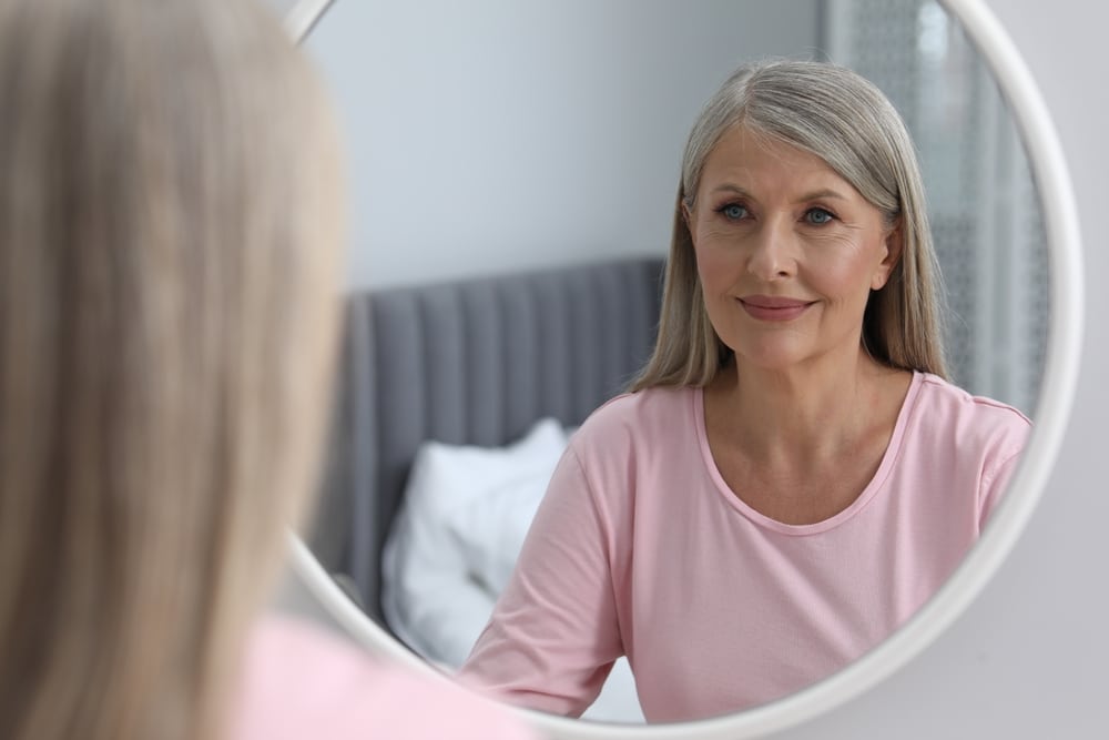 Beauté senior : Femme mûre souriante devant son miroir Femme mûre aux cheveux gris souriant à son reflet dans un miroir rond, vêtue d'un t-shirt rose.